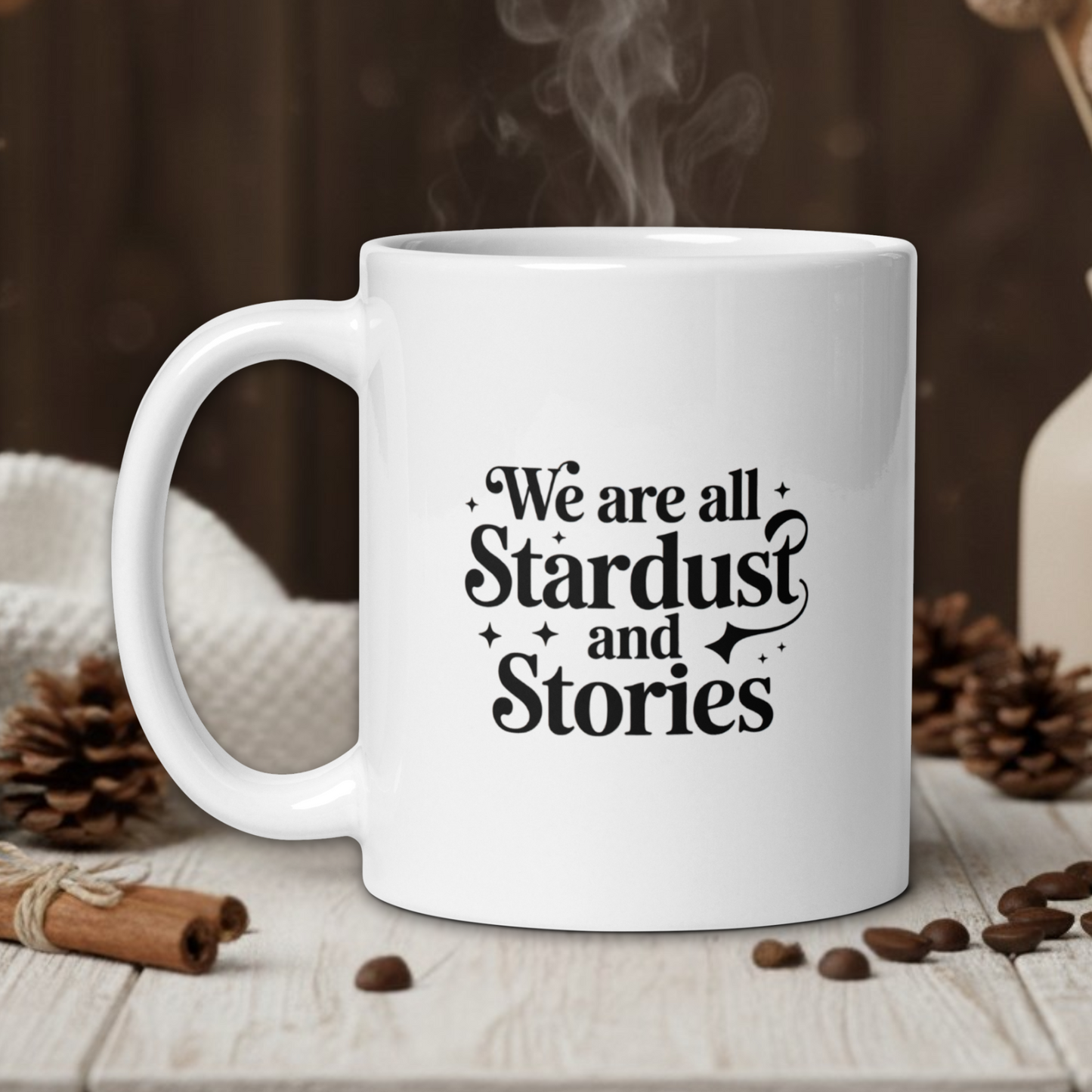 White mug with 'We are all Stardust and Stories' text on a wooden table with coffee beans and a vase in the background.