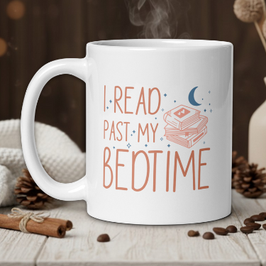 White mug with 'I Read Past My Bedtime' text on a wooden table with coffee beans and a vase.