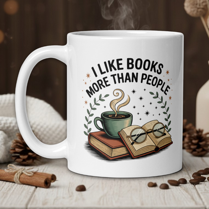 White mug with 'I like books more than people' design on a wooden table with coffee beans and a vase in the background.