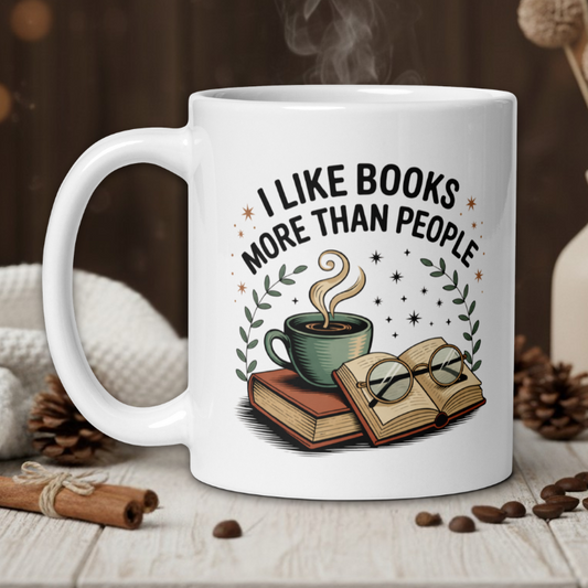 White mug with 'I like books more than people' design on a wooden table with coffee beans and a vase in the background.