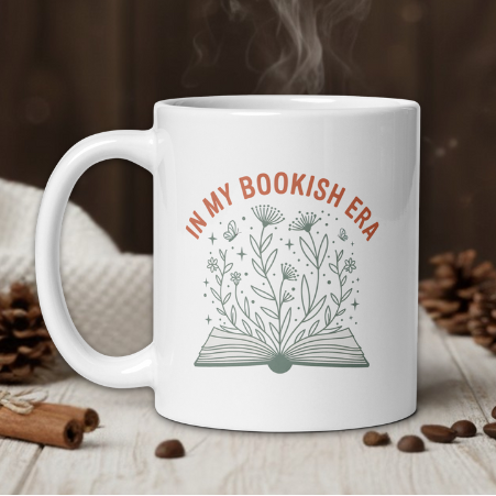 White mug with 'In My Bookish Era' design on a wooden surface with coffee beans and pine cones.