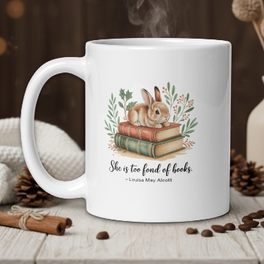 White mug with a rabbit illustration on books, surrounded by coffee beans and a vase with dried flowers.