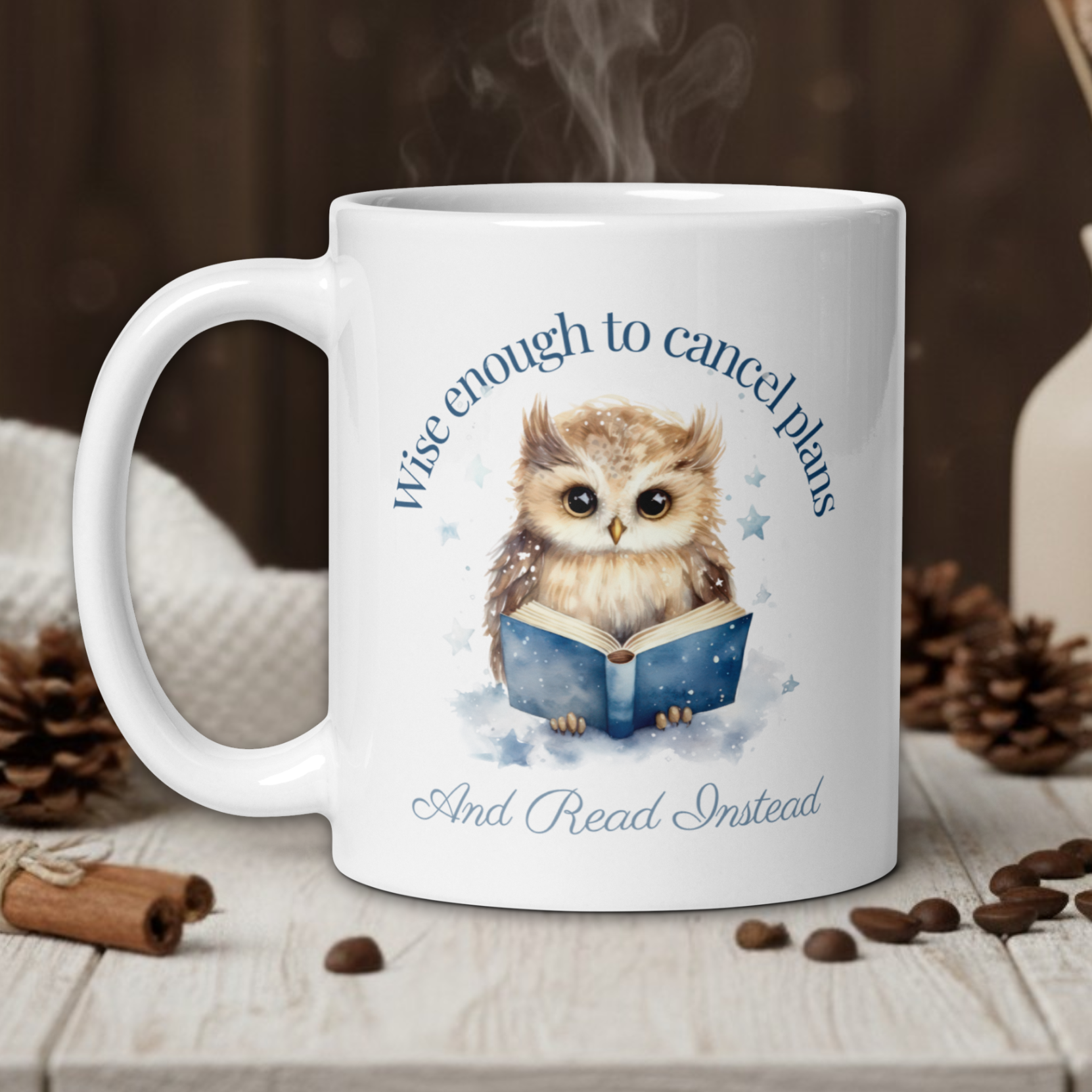 White mug with an owl design and text on a wooden table with coffee beans and a vase in the background.