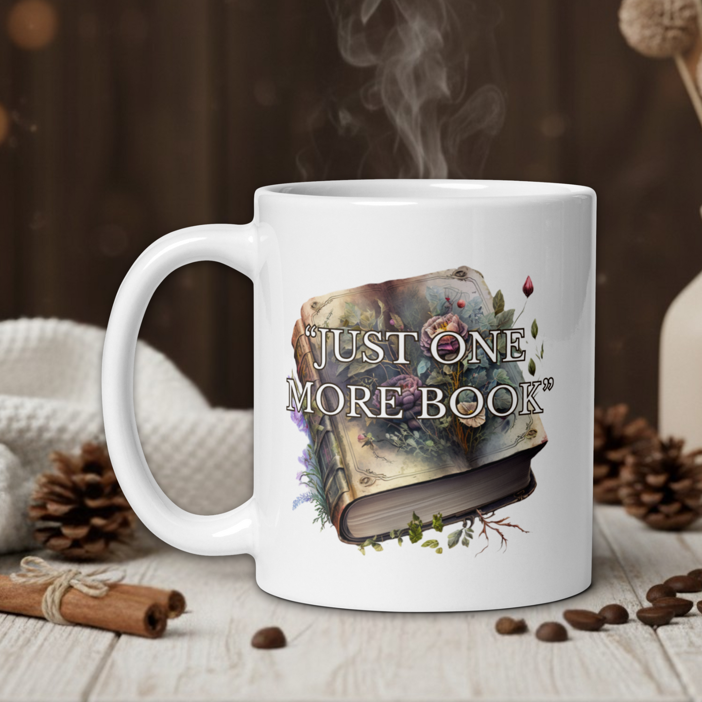 White mug with 'Just One More Book' design on a wooden table with coffee beans and a vase in the background.