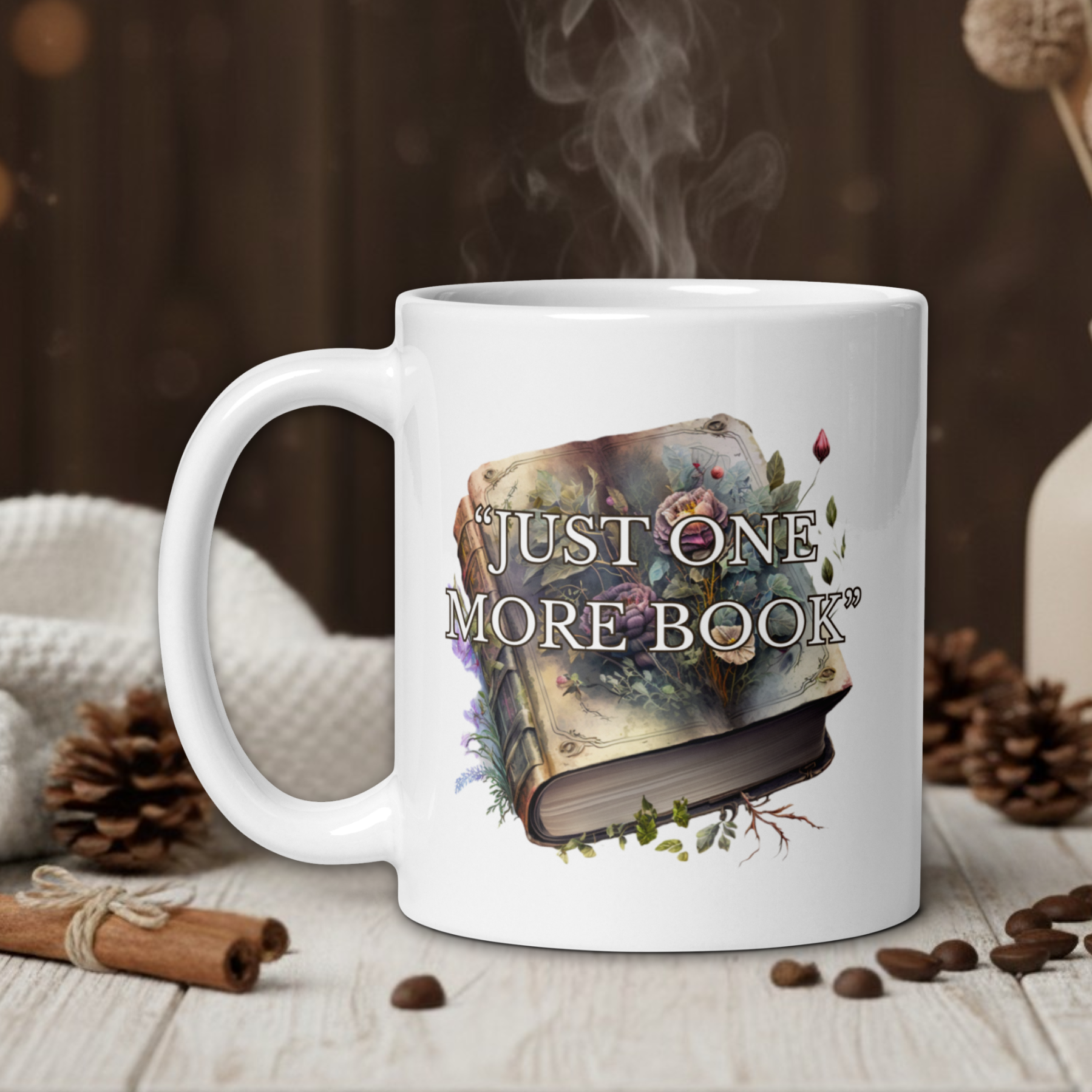White mug with 'Just One More Book' design on a wooden table with coffee beans and a vase in the background.