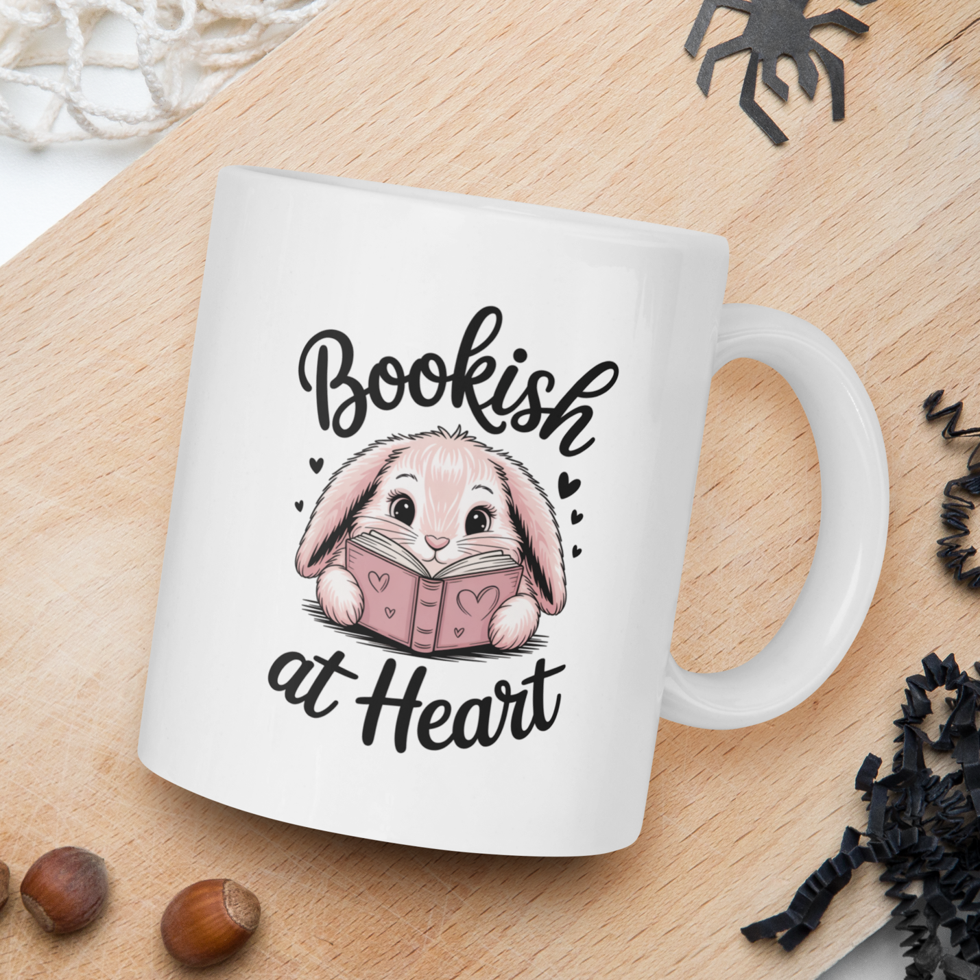 White mug with a rabbit illustration and 'Bookish at Heart' text on a wooden board with nuts and spider decorations.